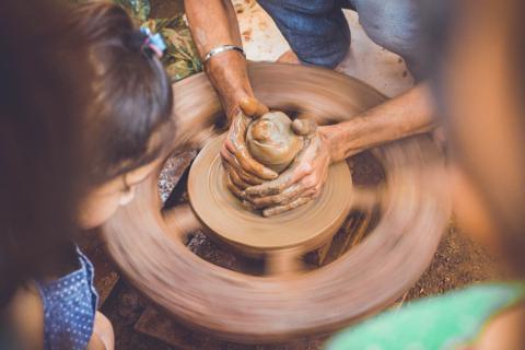Hands cupped around light brown clay in the center of a spinning pottery wheel